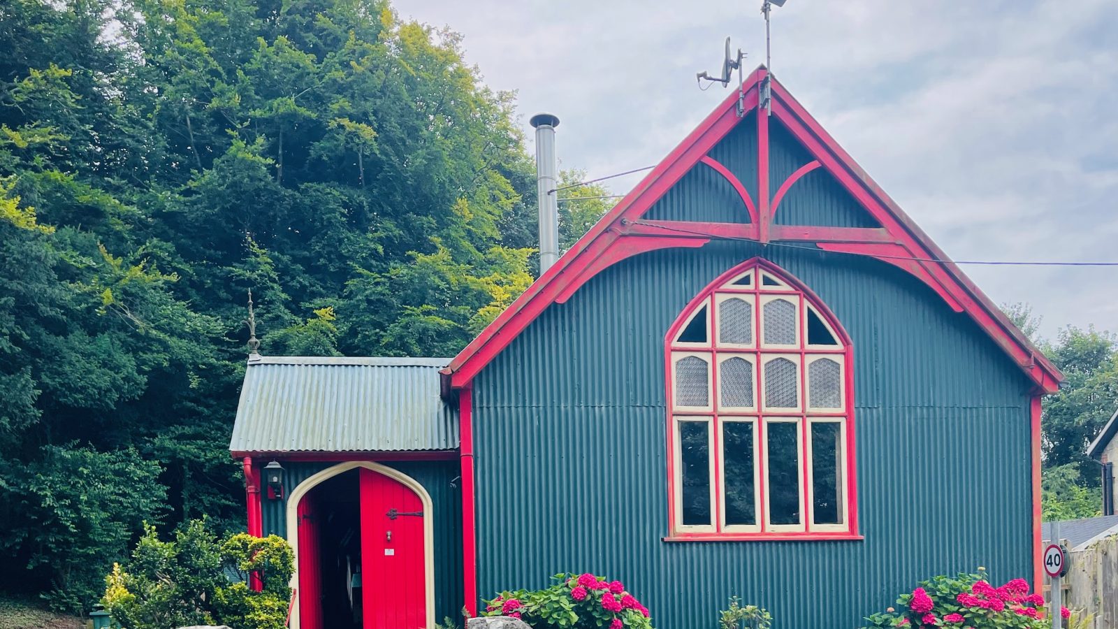 A quaint green house with a red roof and trimmings, featuring arched windows and a chimney, evokes the charm of a Mission-style retreat. Surrounded by vibrant pink flowers and lush greenery, an overcast sky looms while a stone wall marks the front boundary.