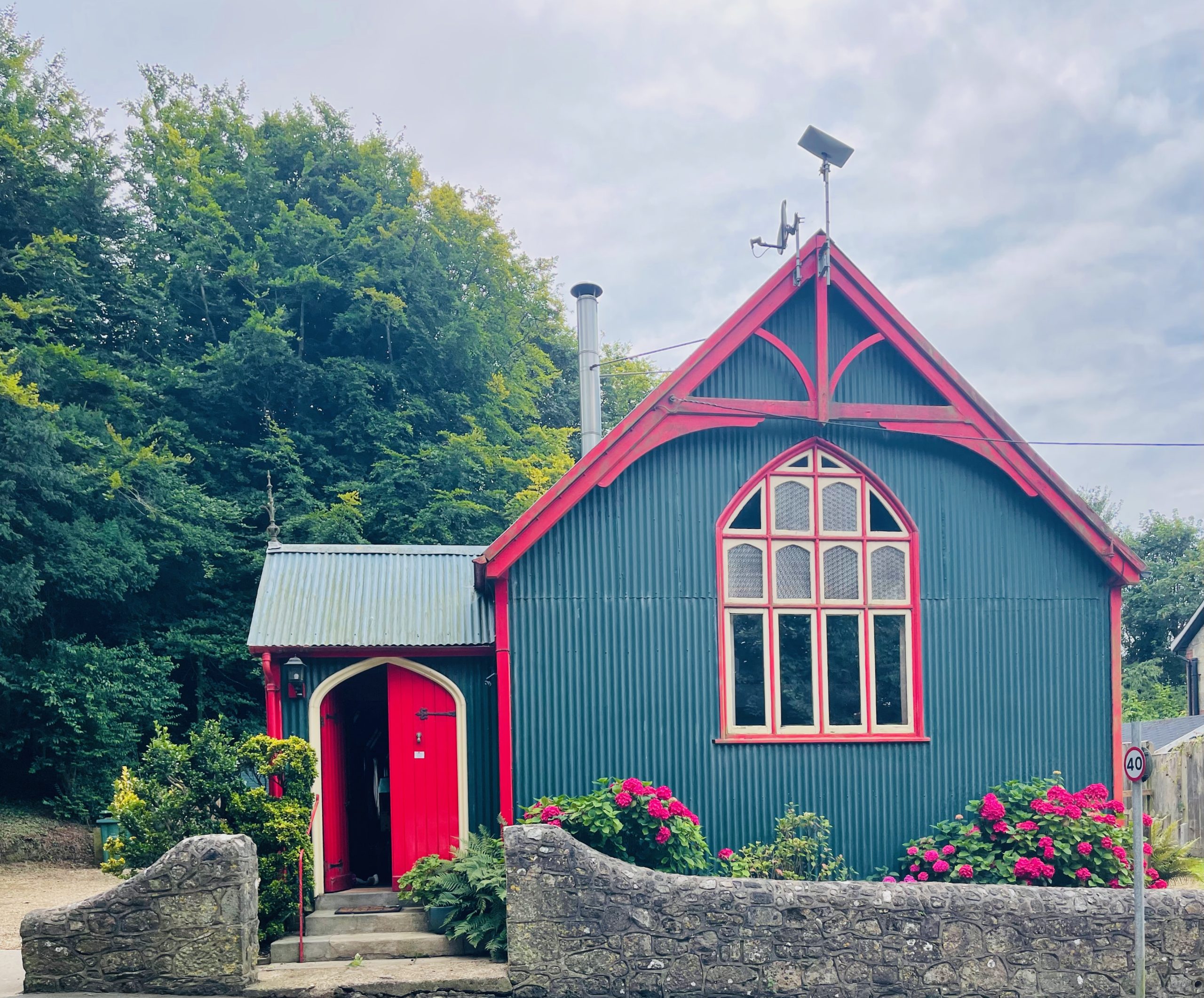 A quaint green house with a red roof and trimmings, featuring arched windows and a chimney, evokes the charm of a Mission-style retreat. Surrounded by vibrant pink flowers and lush greenery, an overcast sky looms while a stone wall marks the front boundary.