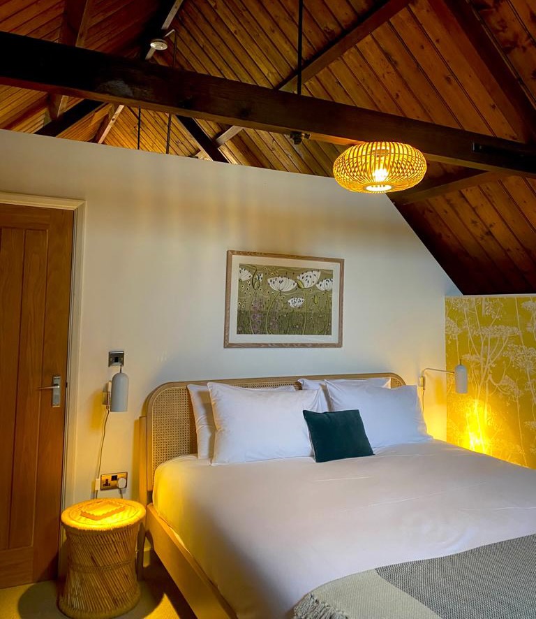 A cozy bedroom with a wooden ceiling evokes The Mission style, featuring a large bed adorned with white linens and a dark green accent pillow. Warm lighting highlights a wicker side table and striking wall art above the headboard, while a wooden door stands slightly open on the left.