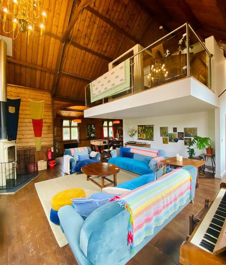 A cozy living room with vaulted wooden ceilings and a mezzanine evokes the charm of The Mission. It features blue sofas, colorful throws, a wooden table, piano, and fireplace. Large windows brighten the space with natural light as a chandelier hang gracefully from above.