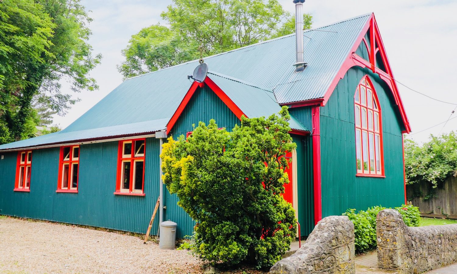 A vibrant teal Mission-style house with red trim and a sloped roof stands amidst lush greenery. It features large windows and a chimney, with a gravel driveway and a charming stone wall at the front.