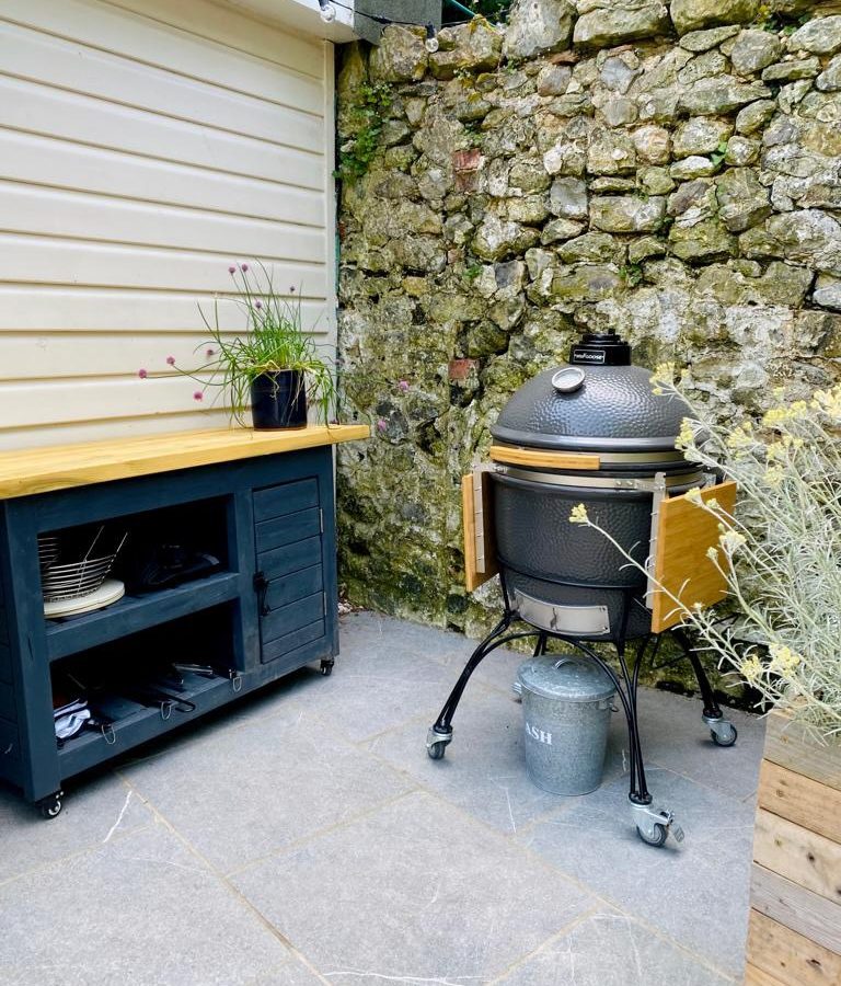 A kamado grill on wheels is positioned on a stone patio next to a rustic stone wall, setting the mission for perfect outdoor cooking. A wooden table with storage shelves is on the left, holding a plant and kitchenware. Nearby, there is a potted plant and a metal ash bin.