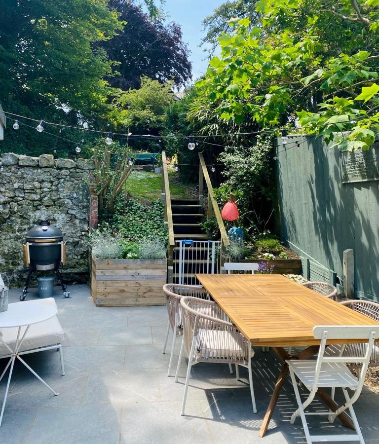 A cozy outdoor patio with a wooden dining table surrounded by chairs sets the stage for gathering. A BBQ grill is on standby, ready for a culinary mission. Steps lead up to a lush garden with a wooden planter, decorative lights strung above as sunlight filters through the trees.