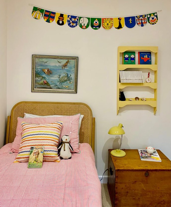 A cozy children's bedroom with a single bed featuring red and white striped bedding and colorful pillows. A stuffed toy sits on the bed, ready for a bedtime mission. The wall is dressed in colorful bunting and art. A yellow lamp and bookshelf sit nearby, while a wooden chest awaits adventures at the foot of the bed.