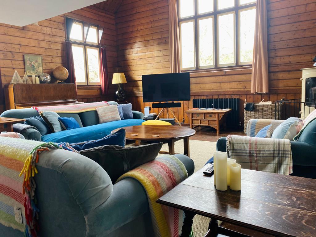 A cozy living room with wooden walls, large windows, and colorful furniture sets the scene for relaxation. A flat-screen TV stands on a tripod in The Mission style, surrounded by sofas and armchairs with blankets. Candles sit on a wooden table next to a piano in the corner.