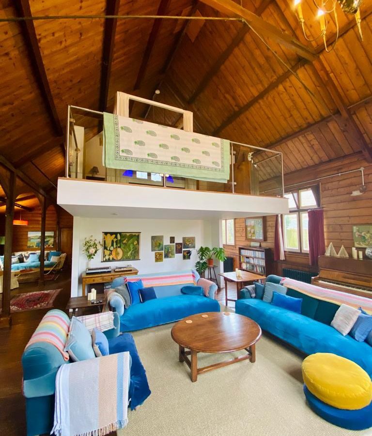 A spacious, cozy living room embodies a Mission style with wooden ceilings, blue velvet couches, and a round wooden coffee table on a light rug. A quilt hangs by an indoor balcony, while paintings decorate the walls. Large windows flood the room with natural light.