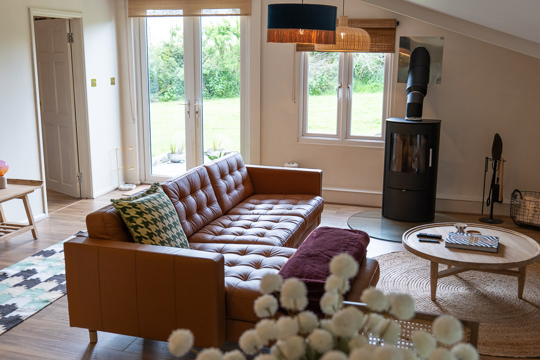 A cozy living room nook with a brown leather sofa, round wooden coffee table, and a black wood-burning stove. Large windows and glass doors open to a garden. Soft lighting and a vase with flowers add warmth to this inviting space.