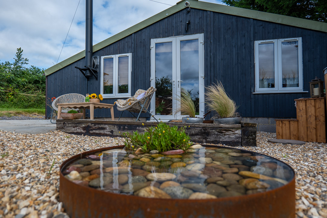 A modern, dark wooden cabin features large glass doors and windows, nestled beside a gravel pathway. A cozy nook with wicker chairs surrounds a table adorned with sunflowers, while a rusted metal water feature filled with stones adds charm to the serene setting.