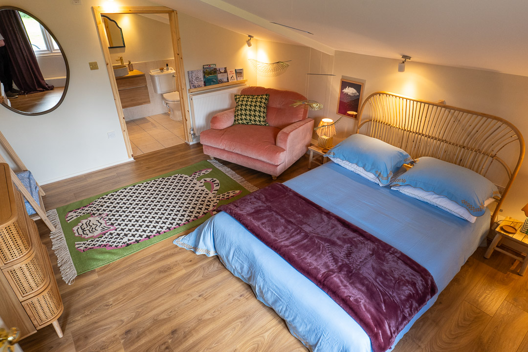 A cozy attic nook reveals a bedroom with a double bed draped in blue sheets and a maroon throw. To the right, a pink sofa nestles near a lamp, completing the inviting space. A green rug with a fish pattern adorns the wooden floor, leading to an open en-suite bathroom in the background.