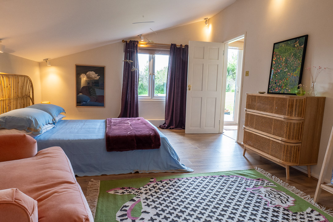 A cozy bedroom, known as The Nook, with a sloped ceiling features a bed adorned with blue and burgundy bedding, a light brown dresser, and a pink sofa. A large window with purple curtains invites natural light, while a colorful rug adds charm to the wooden floor.