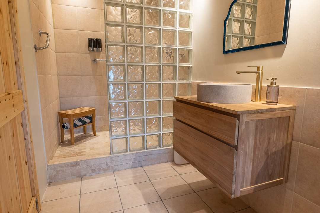 A sleek, modern bathroom features a cozy nook with a wooden vanity, round stone sink, and brass faucet. A glass block shower wall encloses a wooden stool inside the shower area. Beige tiles cover the floors and walls, while a mirror elegantly hangs above the sink.