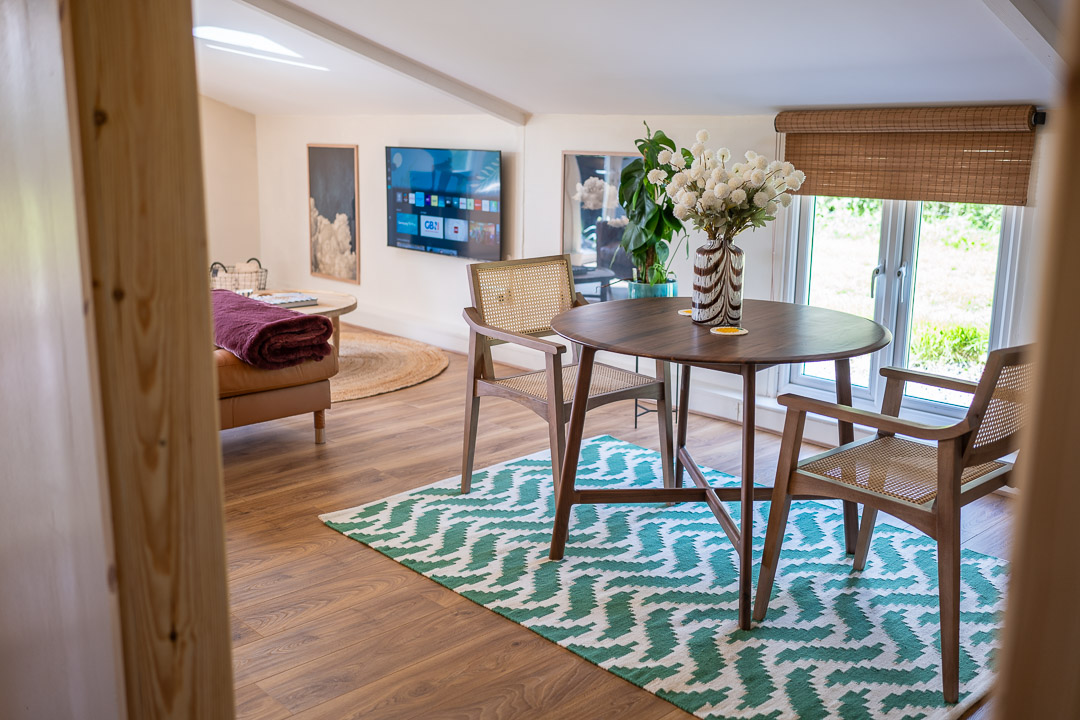 A cozy living space features a wooden table and two chairs atop a teal geometric rug. A brown sofa draped with a purple blanket sits by large windows adorned with bamboo blinds, creating an inviting ambiance. A wall-mounted TV and framed art pieces complete the room, adding character and context.