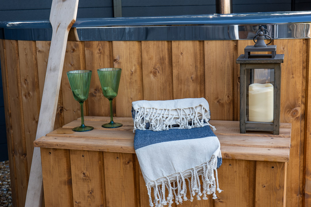 Nestled in the cozy nook of your backyard, this wooden hot tub features a charming wooden step adorned with two green glass goblets, a folded blue and white striped towel, and a lantern with a candle. A rustic ladder leans against the tub, set against a contrasting dark background.