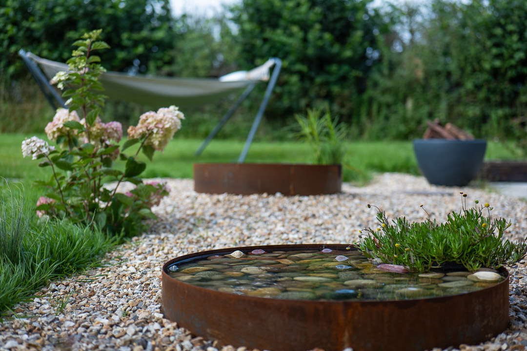 In a serene garden nook, a hammock sways gently amidst lush greenery. In the foreground, a round pond with plants and stones adorns the gravel path, while a bush of pink and white flowers adds vibrant color to the tranquil scene.