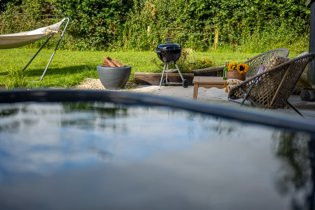 A cozy backyard nook features a hammock, a charcoal grill, and wicker chairs surrounding a low wooden table on lush grass. A potted plant and bouquet of sunflowers brighten the patio, while in the foreground, the pool edge mirrors the vast sky above.