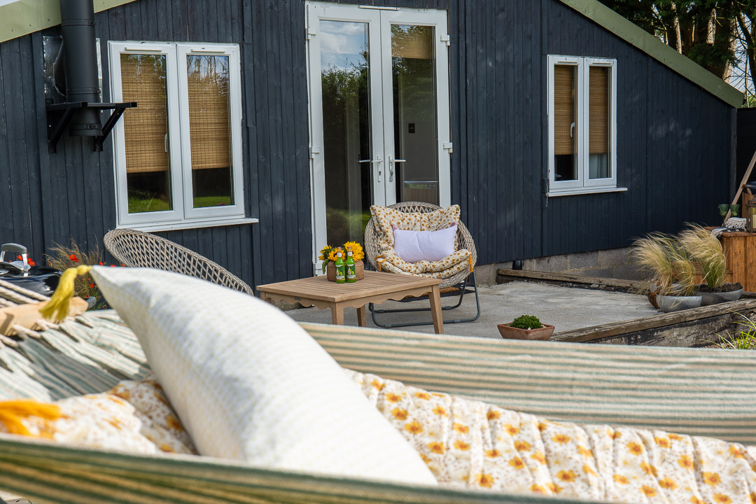 A cozy space featuring an outdoor patio with wicker chairs and a wooden table adorned with flowers. A striped hammock with a pillow invites relaxation in this tranquil garden nook, set against the backdrop of a dark wooden house with two windows and a welcoming door.