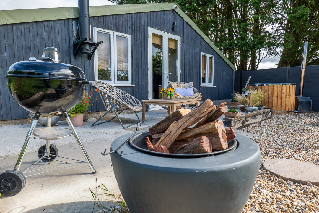 A cozy outdoor patio called The Nook features a fire pit filled with logs in the foreground and a charcoal grill nearby. A modern wooden house with large windows, surrounded by greenery, provides inviting outdoor seating in the background.