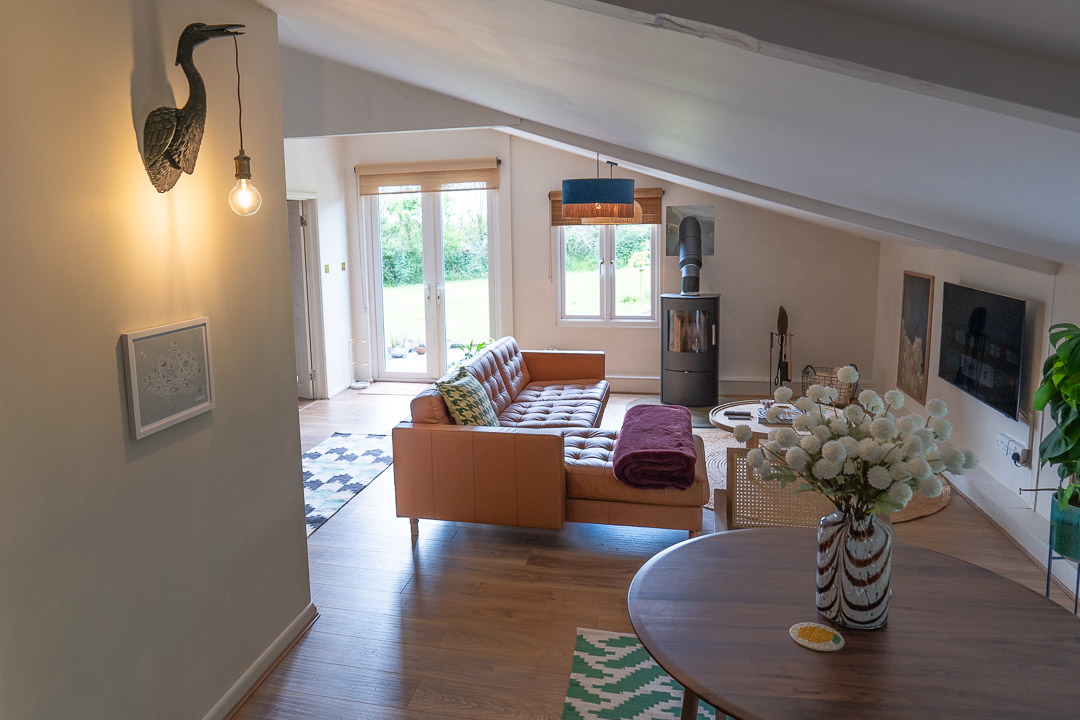 A cozy living room nook with a brown leather couch, a wood-burning stove, and a large window overlooking the garden. The space features wooden flooring, a round table with a vase of white flowers, and modern lighting. A heron-shaped wall lamp adds charm to the inviting corner.