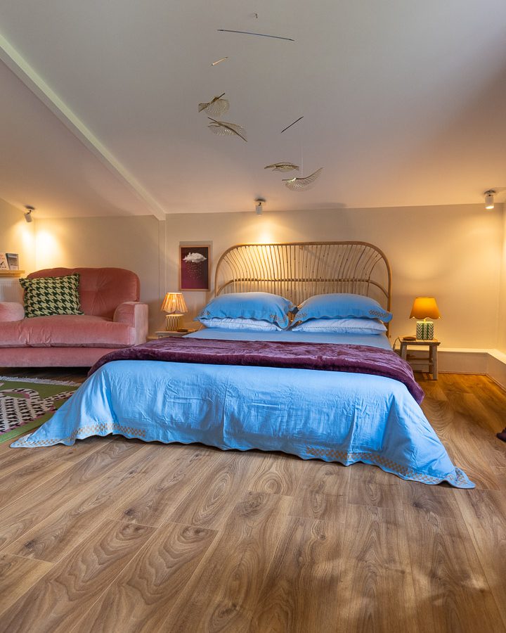 A cozy bedroom nook with a wooden floor features a double bed adorned with blue bedding. A rattan headboard complements the modern decor. On one side, a pink sofa and a small table lamp add charm, while artistic mobiles subtly hang from the ceiling.