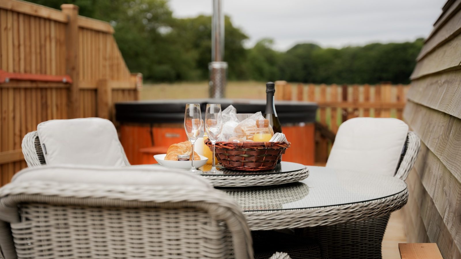 A wicker patio table with four chairs sits on the wooden deck at Partridge Retreat. The table is set with champagne glasses, a wine bottle, and a wicker basket of snacks. In the background, there's a wooden fence and hot tub. Trees can be seen in the distance, enhancing this tranquil escape.