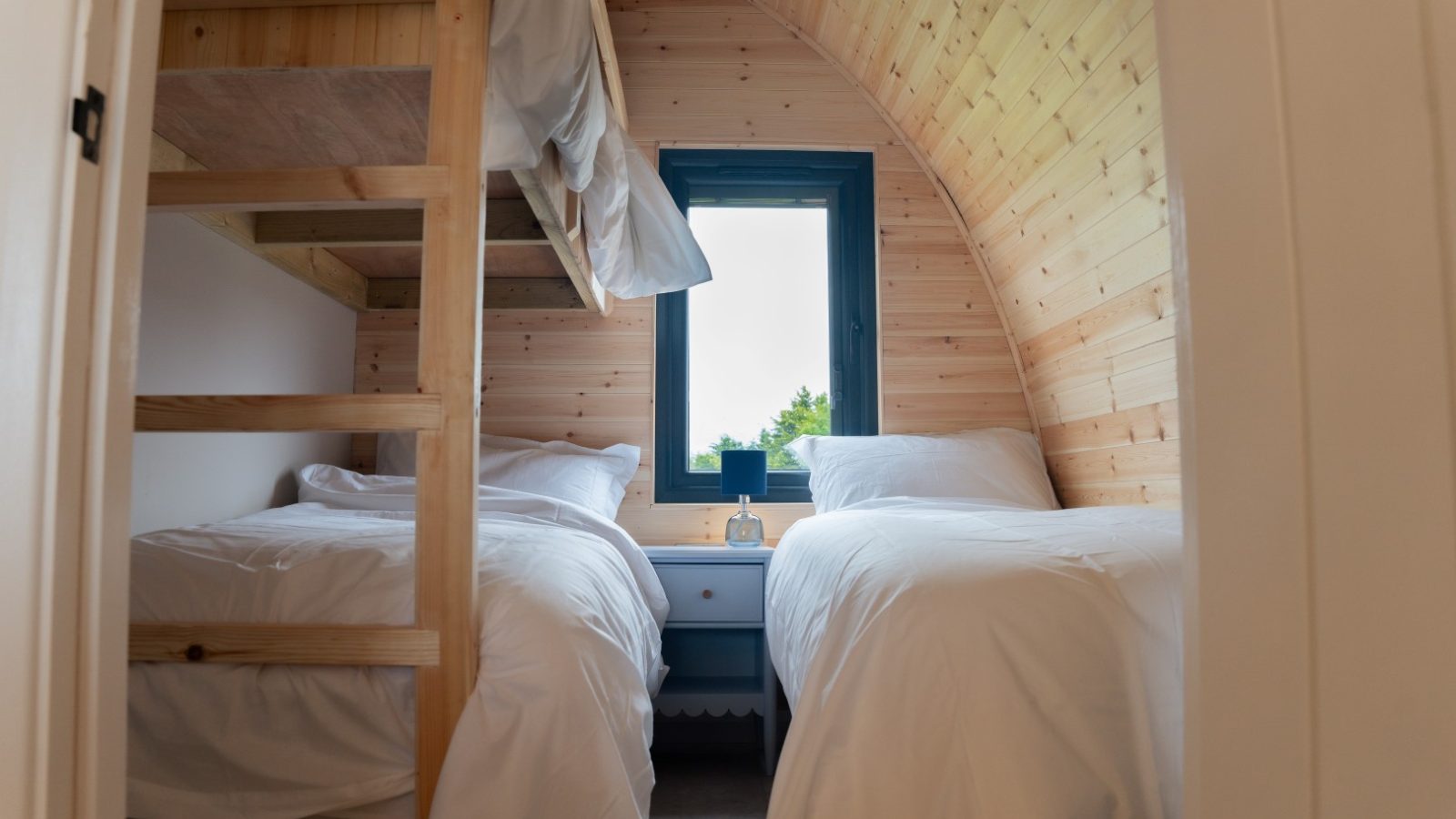 The Partridge Retreat's cozy cabin bedroom boasts two white twin beds and a small window in between. With wooden walls and a bunk bed on the left, it creates a warm, rustic atmosphere that embodies the charm of this serene escape.