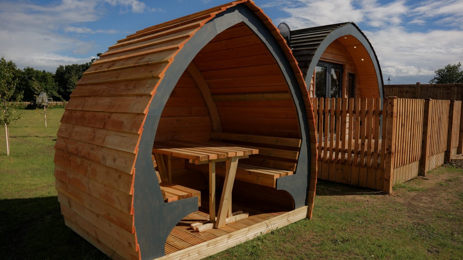 Wooden, pod-shaped cabins at Partridge Retreat stand on a grassy field under a blue sky. One cabin features an open seating area with a table and benches. A wooden fence partially encloses this serene retreat space, perfect for relaxation and nature enjoyment.