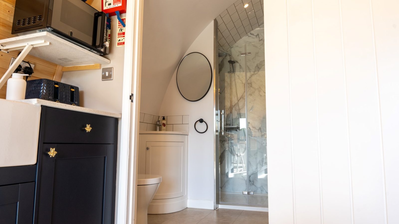 Discover the Partridge Retreat's cozy bathroom featuring a round mirror above a sleek sink, a modern shower with glass doors, and a pristine white toilet. The adjacent kitchenette offers convenience with its microwave, shelves, and cupboard.