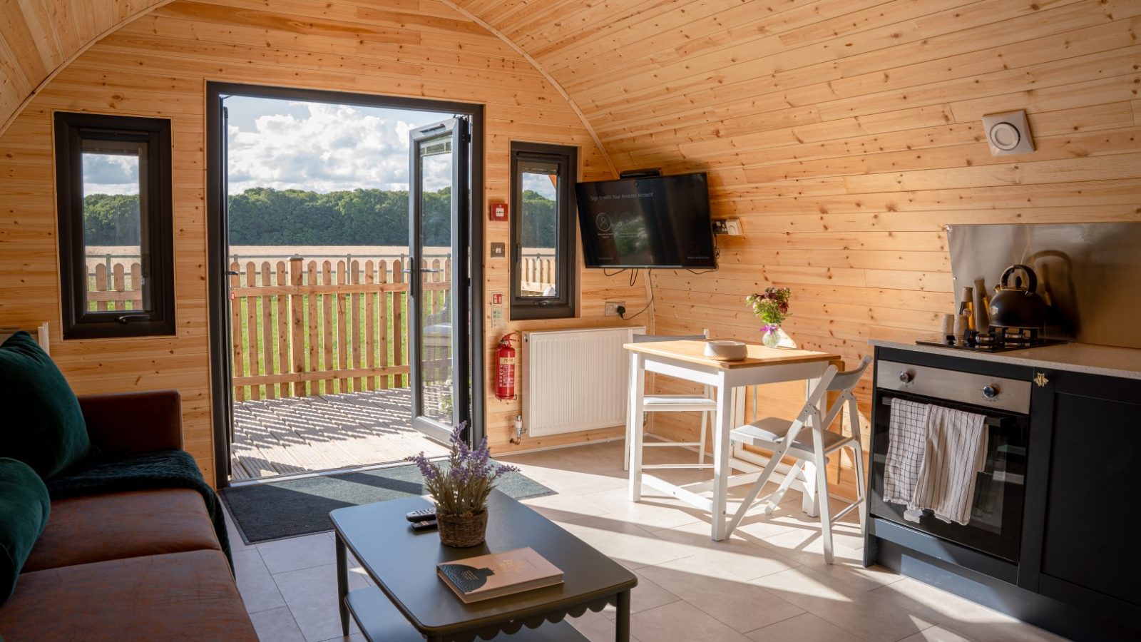 The cozy wooden cabin interior, dubbed Partridge Retreat, boasts an arched ceiling, a comfy sofa, and a coffee table with a vase. A TV and dining table complement the kitchenette. Double doors lead to a deck with scenic views of the field and trees bathed in natural light.