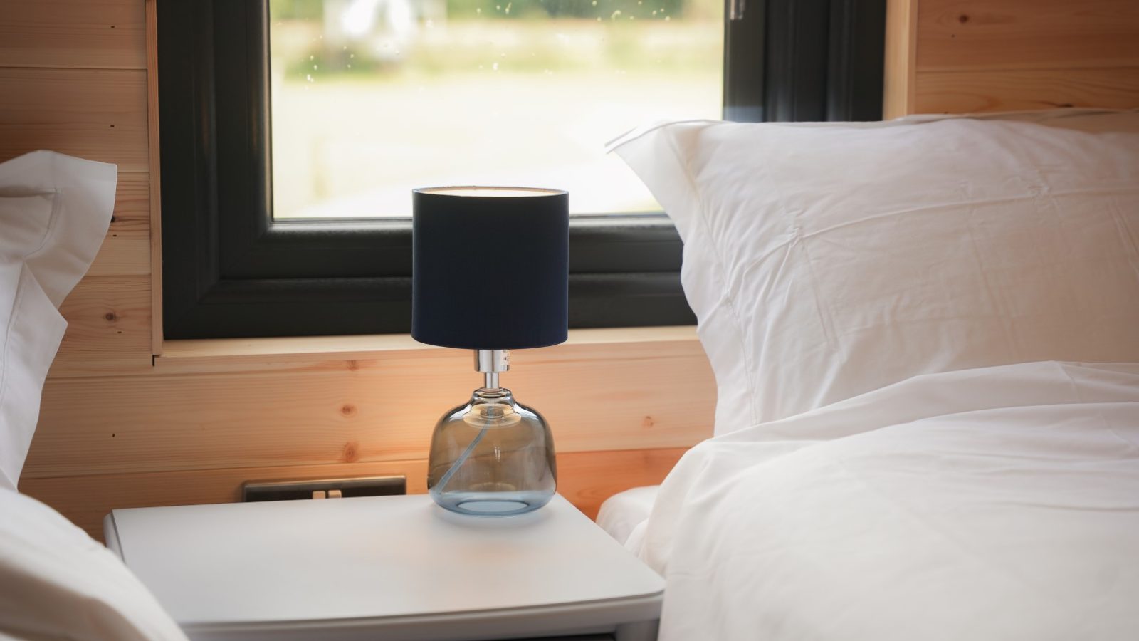 A cozy bedroom scene at Partridge Retreat features two beds with white linens. A small blue lamp rests on a white nightstand between them. Behind the beds, a window frames a blurred outdoor view, highlighting the warm, natural wood-paneled wall that defines this serene getaway.