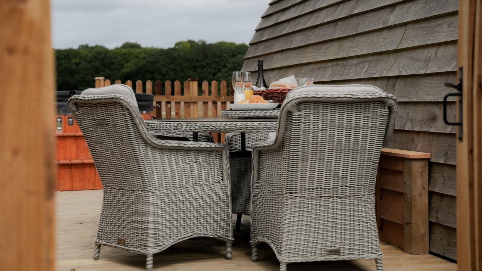 A cozy outdoor setting at Partridge Retreat features a round wicker table with two matching chairs on a wooden deck. The table holds a glass of wine, a bottle, and sliced bread. A curved wooden structure and lush greenery add to the charming ambiance in the background.