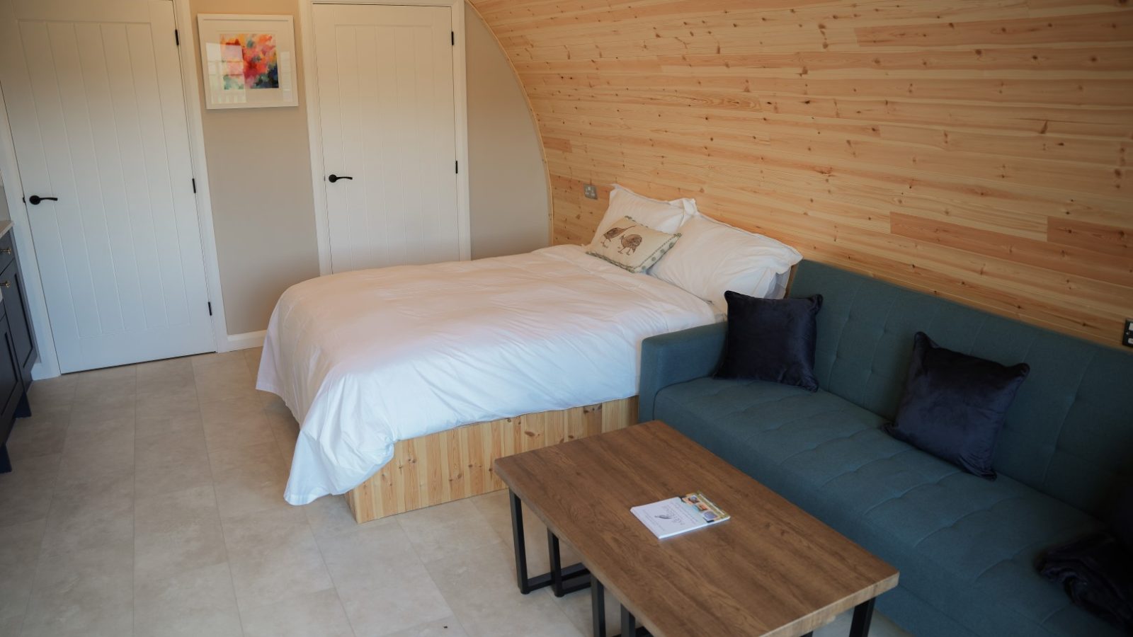 A cozy room at Partridge Retreat boasts a curved wooden ceiling, a bed with white linens, and a teal sofa with dark cushions. A wooden coffee table holds a magazine, while framed artwork adorns the light-colored wall beside two white doors.