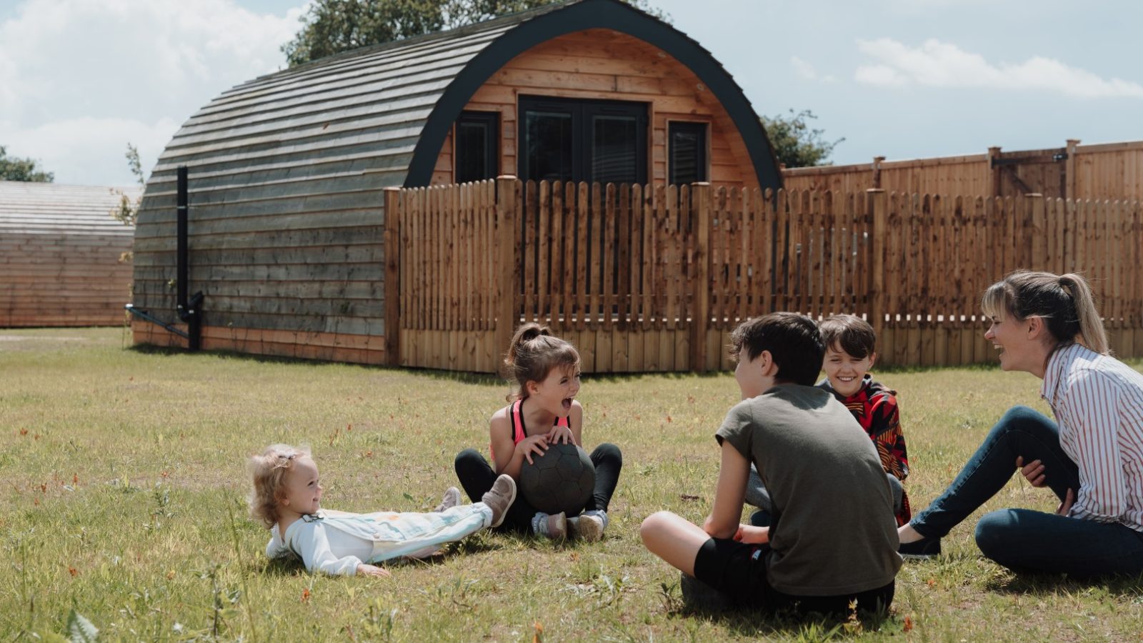 A group of children and an adult sit on the grass in a sunny field at Partridge Retreat, laughing and talking. Behind them are wooden, pod-like cabins and a wooden fence. The sky is clear with some clouds above.