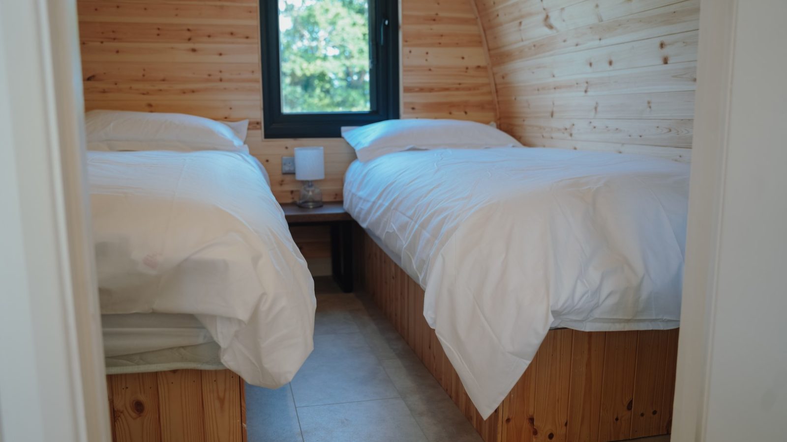 Discover a cozy bedroom at Partridge Retreat, featuring two single beds with white linens. Light-colored wooden walls and bed frames complement the space. A window between the beds offers natural light and views of green trees, while a small lamp rests on a corner shelf for your comfort.