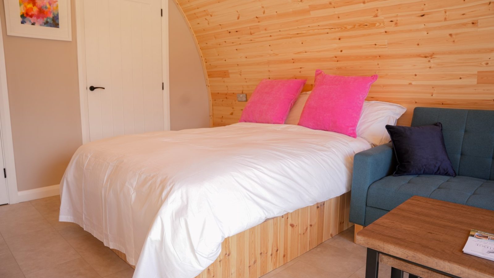 Nestled in the Partridge Retreat, this cozy bedroom boasts a wood-paneled arch ceiling, a bed with white linens and pink pillows. A teal sofa with a purple cushion complements the wooden coffee table, while a colorful abstract painting adorns the wall near the white door.