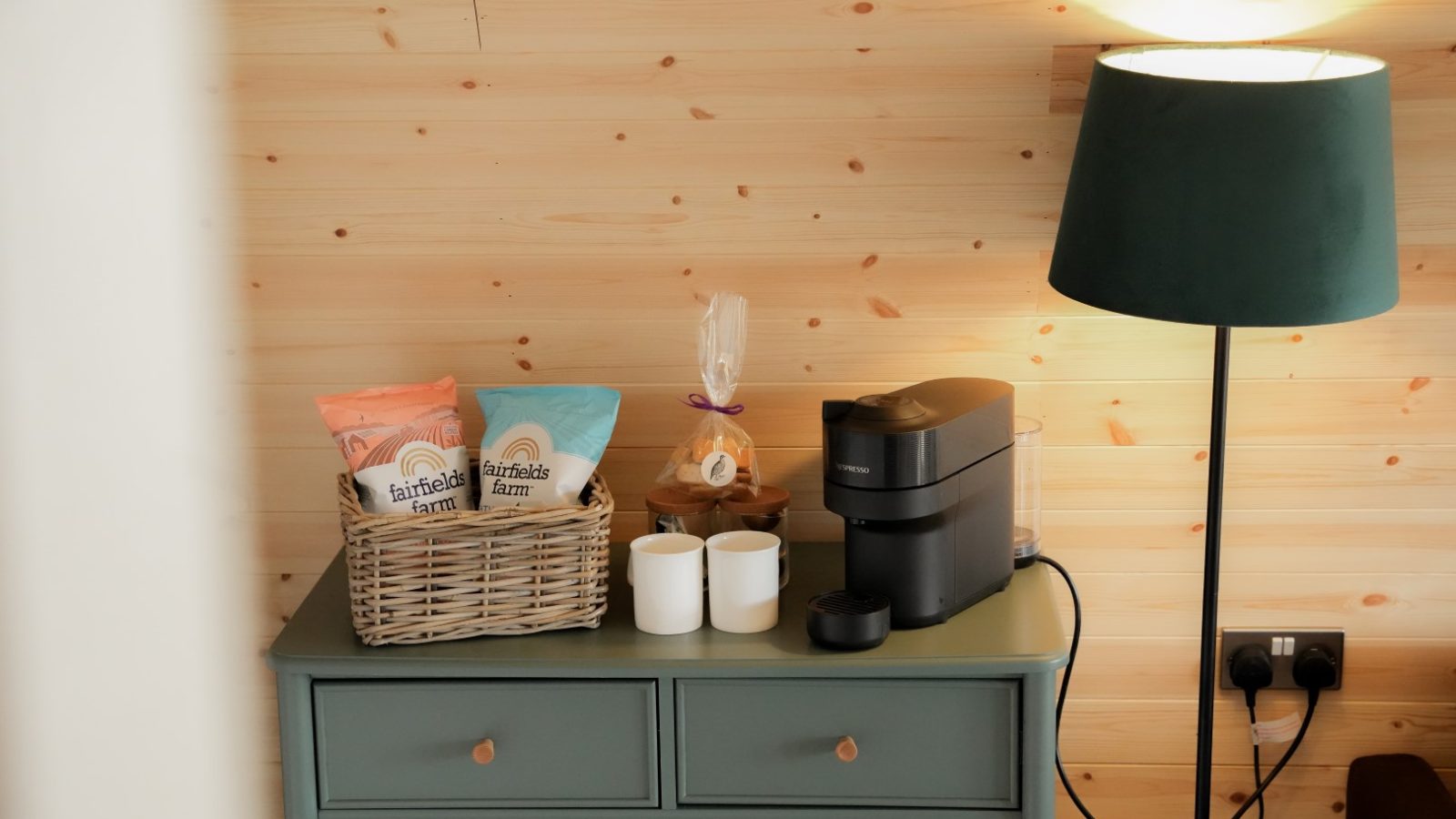 In a cozy corner at Partridge Retreat, a green cabinet holds a coffee machine, two mugs, a basket of snack bags, and a small wrapped gift. A tall green lampshade casts warm light against the wooden wall.