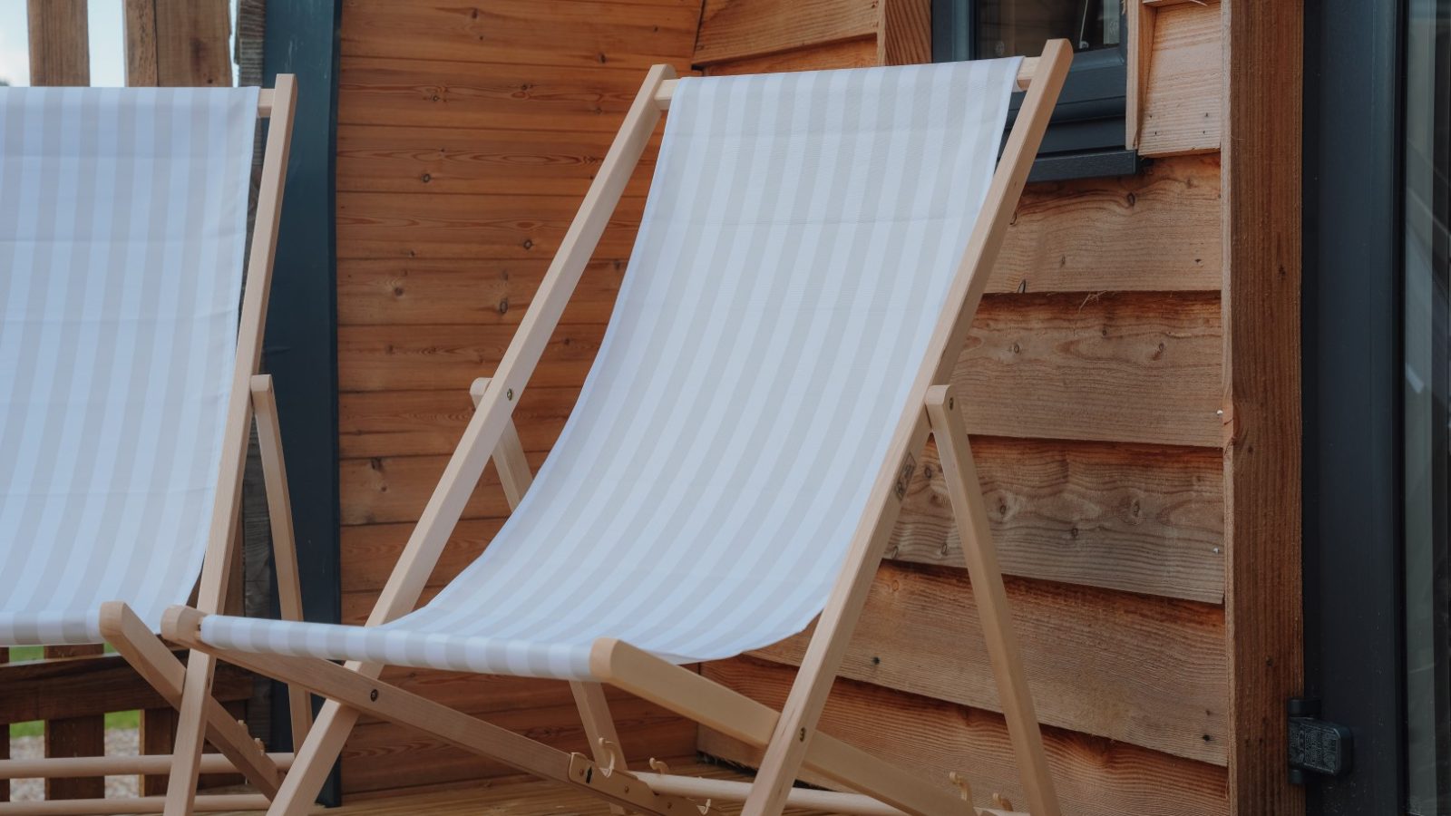 Two wooden deck chairs with white striped fabric are set on a wooden patio, embodying the tranquil charm of Partridge Retreat. A wooden cabin wall with a window enhances this cozy outdoor setting.