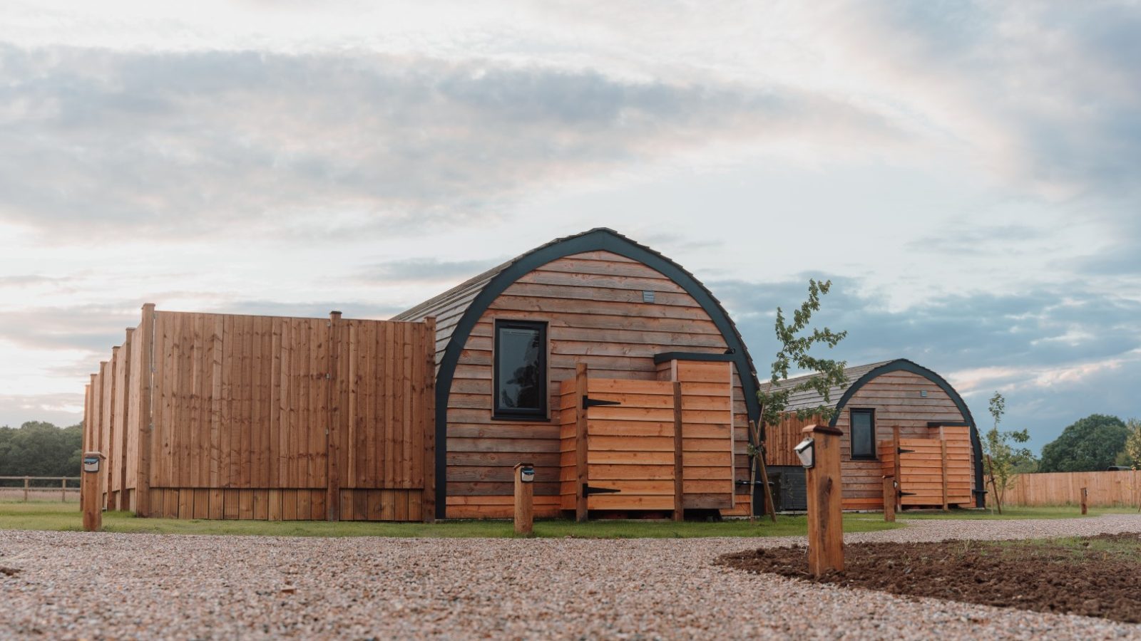 Two wooden glamping pods with arched roofs sit on a gravel path at Partridge Retreat, surrounded by grassy fields and a cloudy sky. Small wooden fences partially enclose each pod, and a few young trees are visible nearby, creating the perfect serene escape.