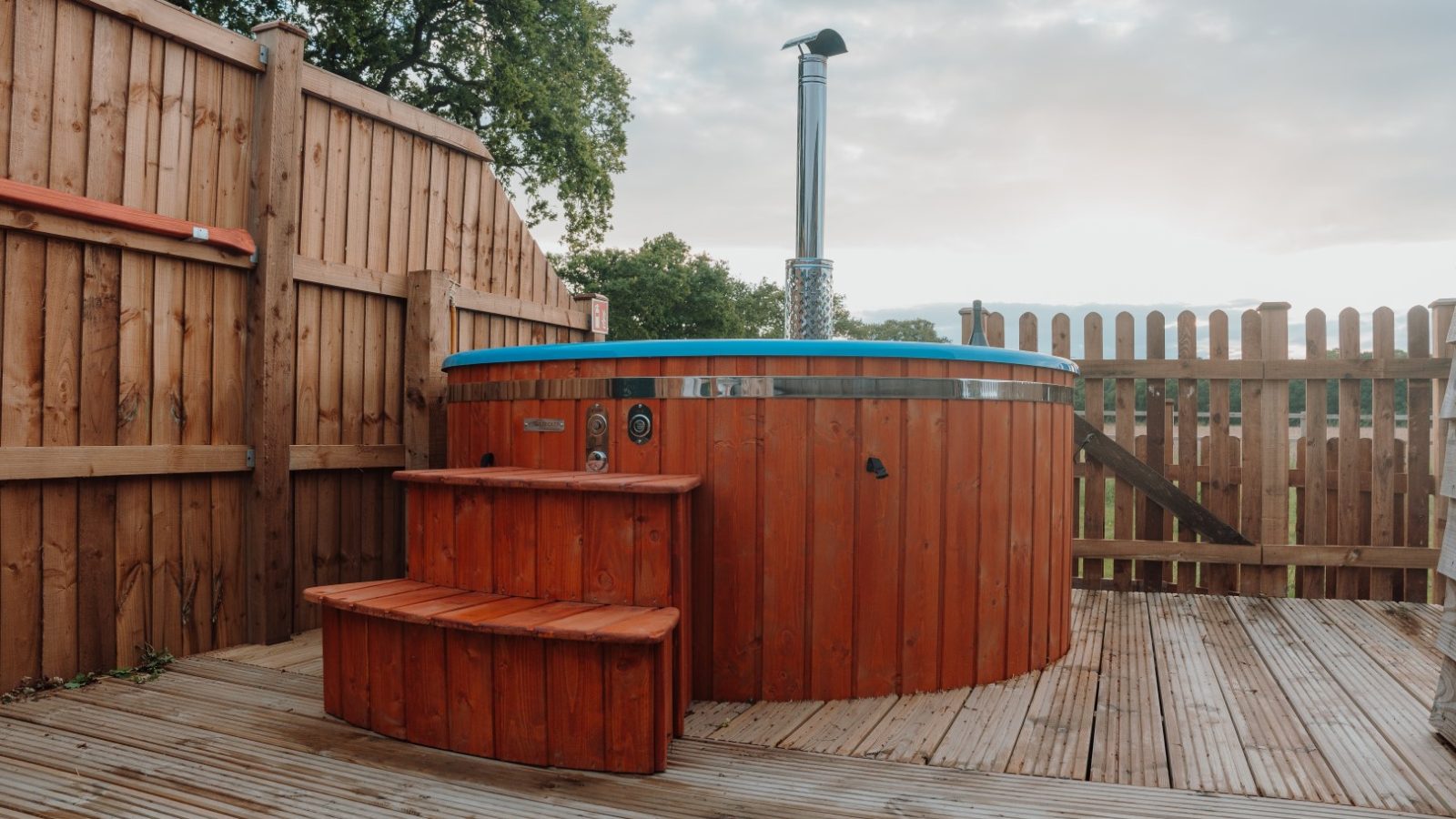 A wooden hot tub with a small set of matching steps is set on a wooden deck at Partridge Retreat, surrounded by a charming wooden fence. The sky is partly cloudy, with trees visible in the background, creating the perfect Partridge Retreat experience.