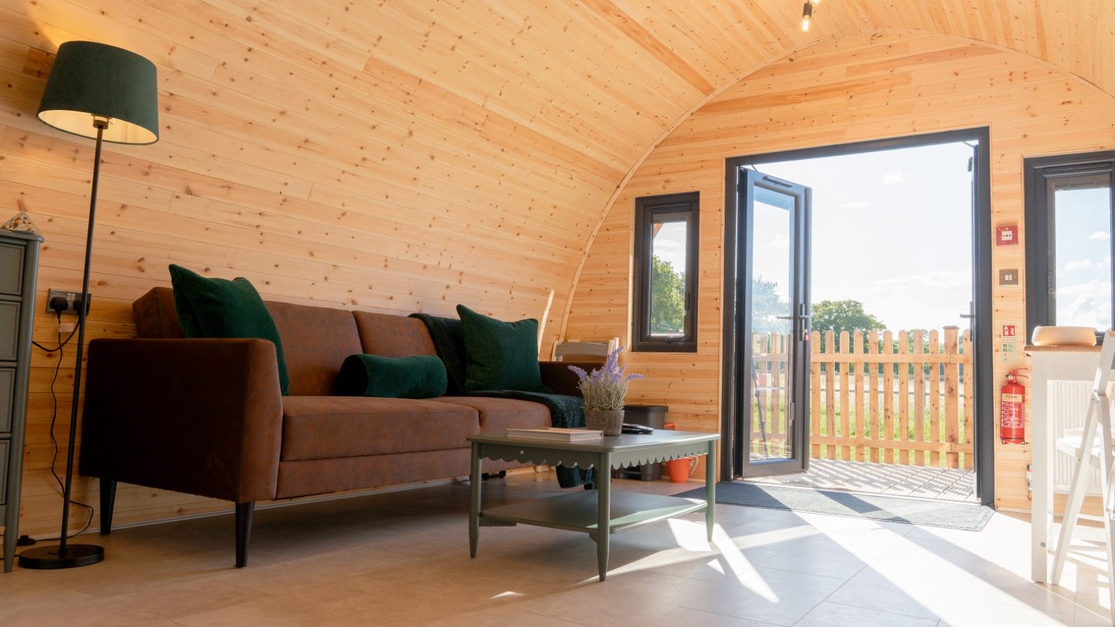 The interior of the cozy wooden Partridge Retreat cabin boasts a curved ceiling. It features a brown sofa with dark green cushions, a coffee table with a small plant, and large windows with an open door leading to a sunny patio offering views of the wooden fence and trees.