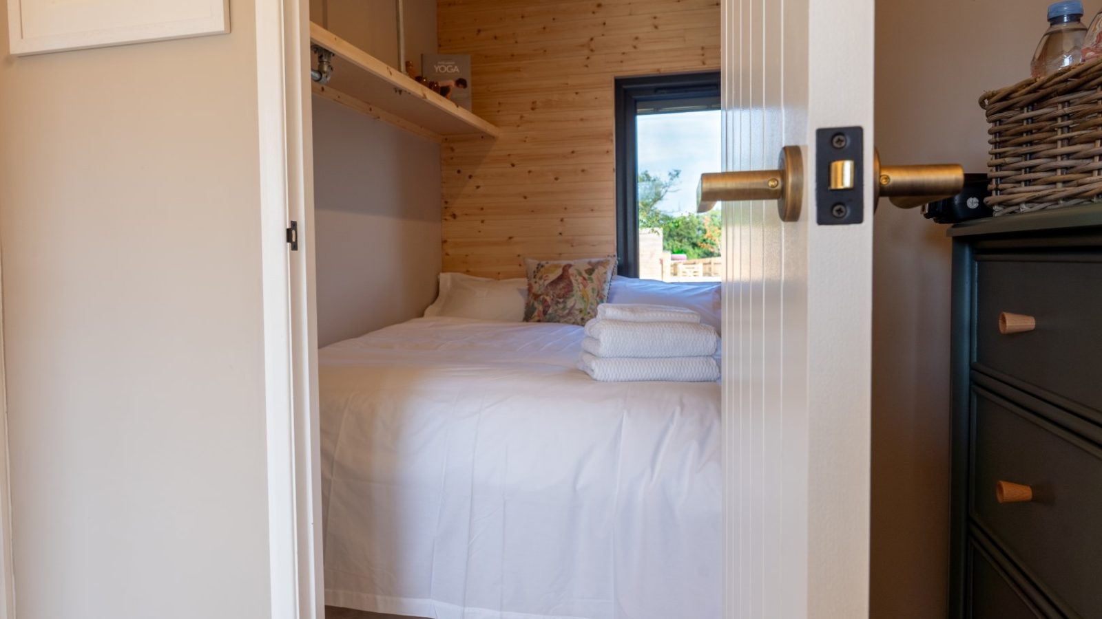 A cozy bedroom at Partridge Retreat features light wood paneling and soft beige walls, visible through an open door. A neatly made bed with white linens, colorful cushions, and folded towels sits by a window. Nearby, a black dresser and a wicker basket complete the tranquil setting.