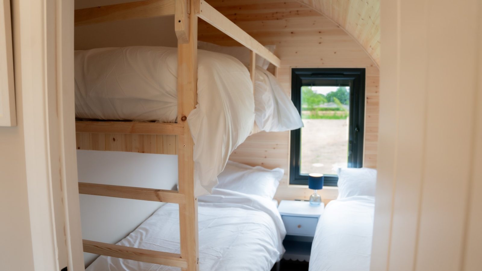 A cozy small bedroom with wooden walls at Partridge Retreat features a bunk bed and a single bed, both adorned with white linens. A quaint bedside table with a lamp sits between the beds, while a window offers an inviting view of the lush greenery outside.