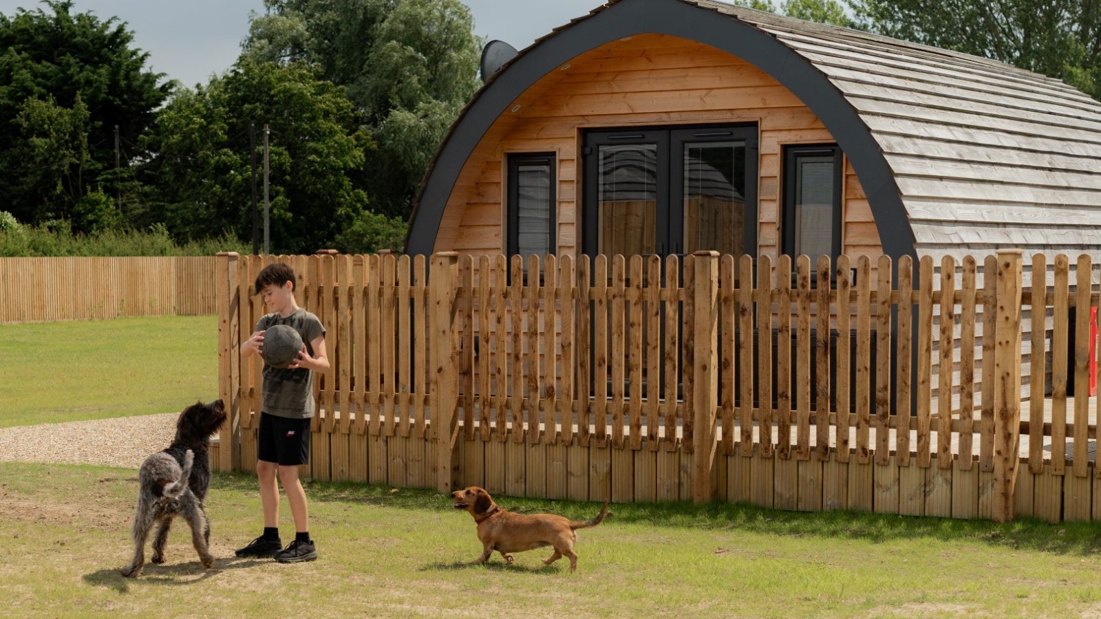 At Partridge Retreat, a child stands in front of a wooden cabin, playing with a large black dog. A small brown dog strolls nearby on the grassy area. A wooden fence surrounds the cabin, framed by trees under a cloudy sky.