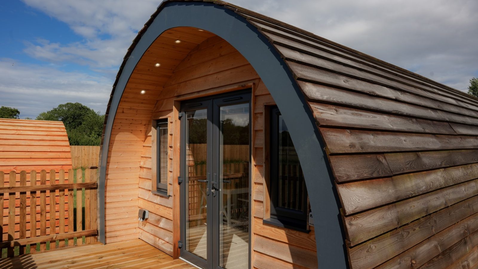 Nestled in the serene embrace of nature, this cozy wooden cabin at Partridge Retreat boasts a curved roof and expansive glass doors, opening onto a spacious deck. The partly cloudy sky casts playful shadows over the wooden fence and surrounding trees, creating an idyllic escape.