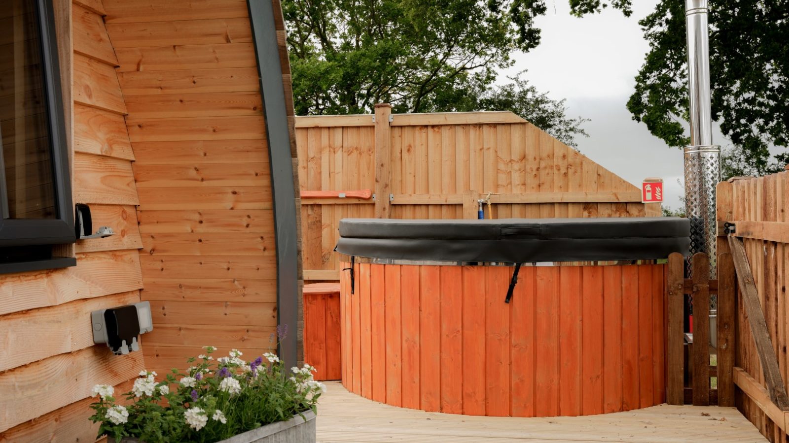 A wooden cabin with a deck features a round hot tub, creating the perfect Partridge Retreat. A metal planter brimming with flowers adorns the deck, while a wooden fence and trees provide a serene backdrop.