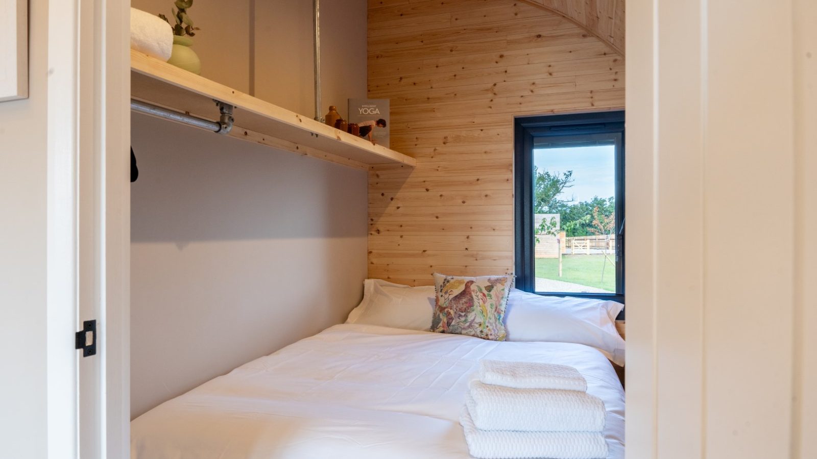 Cozy bedroom with wood-paneled walls and a large window at Partridge Retreat. A neatly made bed with white linens and a colorful pillow is in the foreground. A shelf above holds a rolled towel, small plant, and a book. The window offers a view of greenery outside.