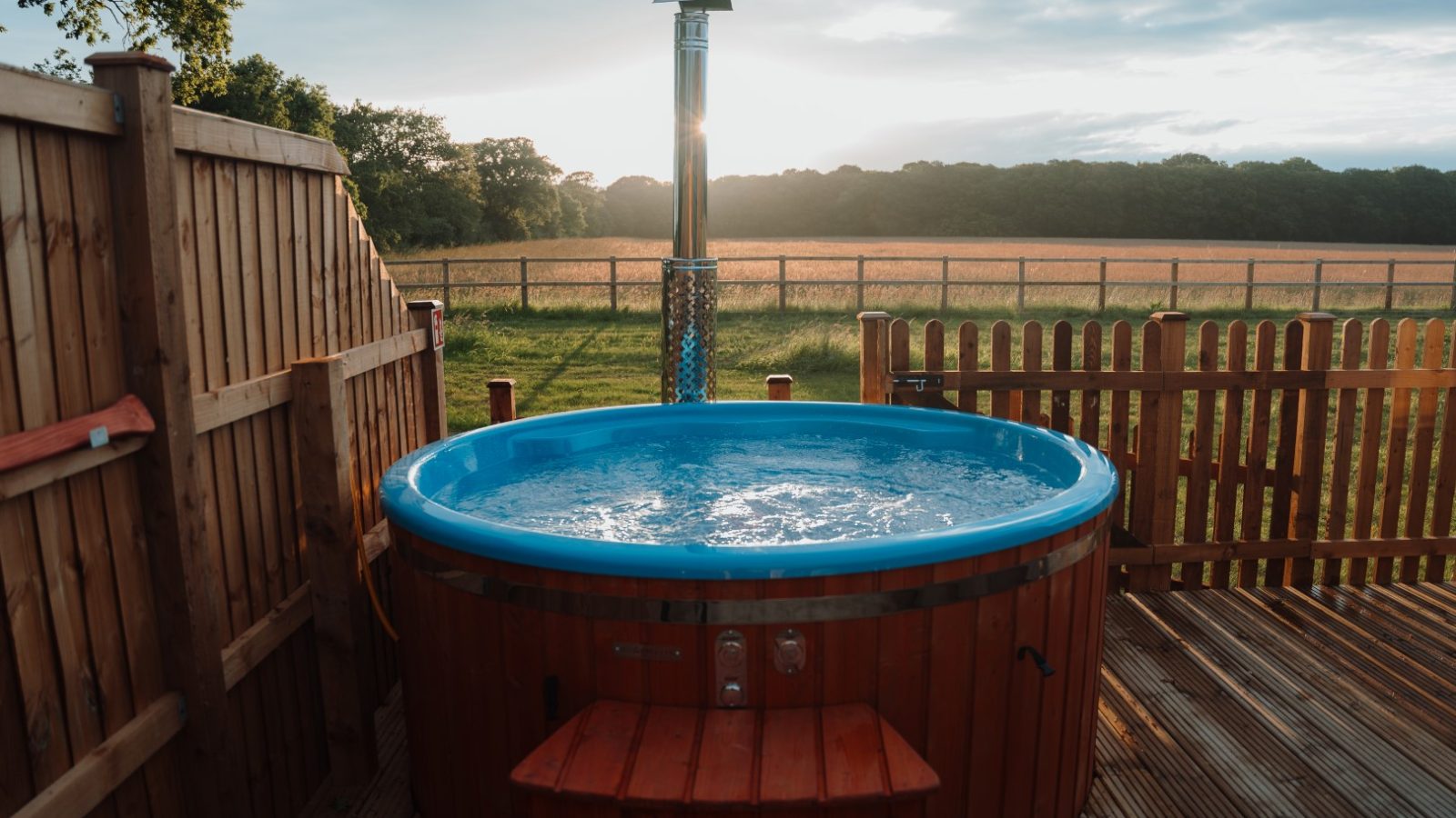 A round hot tub with bubbling water is situated on a wooden deck at Partridge Retreat, enclosed by a fence. Beyond the enclosure, there's a grassy field under a cloudy sky with sunlight peeking through. Trees line the horizon, enhancing the tranquil atmosphere of this retreat.