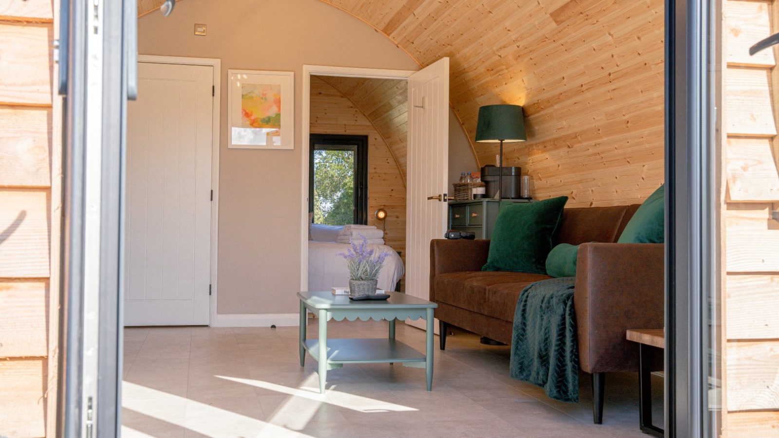 Cozy cabin interior at Partridge Retreat boasts wooden walls, a brown sofa adorned with green pillows, and a small coffee table. A floor lamp stands by the sofa, while framed art decorates the walls. Light streams through an open door leading to a tranquil bedroom.