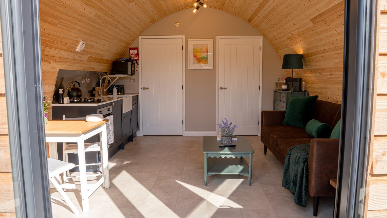 Partridge Retreat offers a cozy, arched wooden interior with a kitchenette, small dining table, and sofa. The room is adorned with a potted plant and wall art. Natural light pours in from a large open door, illuminating the light wood walls and tiled floor for a serene escape.