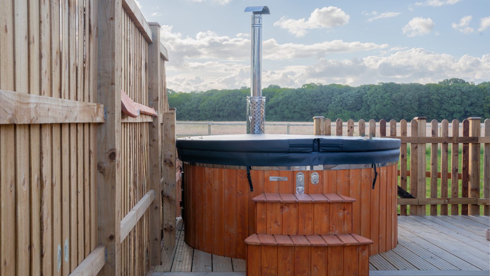 A wooden hot tub with a black cover and metal chimney sits on a wooden deck at the Partridge Retreat. Surrounded by a wooden fence, steps lead up to it. The backdrop features a field and trees under a partly cloudy sky, perfect for an idyllic Partridge Retreat getaway.