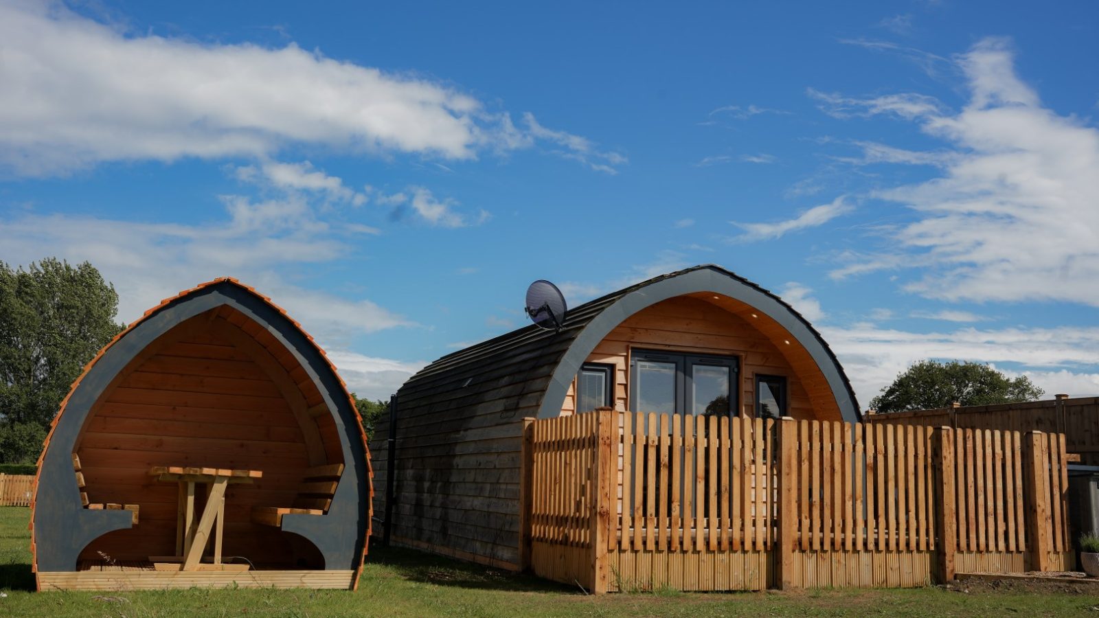 A wooden pod cabin with large windows and a fenced patio stands on a grassy area under a blue sky, perfect for a cozy Partridge Retreat. Next to it is a smaller, open shelter with a picnic table inside. Trees grace the background, adding to the tranquil setting.